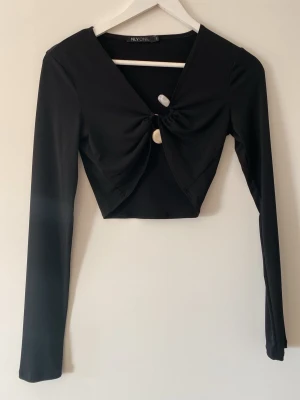 Svart croppad topp från NLY One - Trendig svart croppad topp från NLY One med lång ärm och snygg knutdetalj framtill. Djup v-ringning och figurnära passform gör den perfekt till jeans eller kjol. Tunt och stretchigt material.