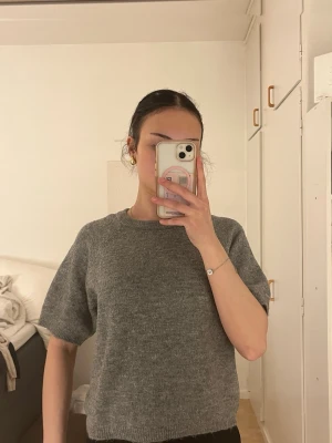 Grå stickad t-shirt i ullmix - Mjuk och enkel grå stickad t-shirt med korta ärmar och rund halsringning. Perfekt för lager-på-lager och har en avslappnad passform. Materialet ser ut att vara en ull- eller mohairmix som ger en mysig känsla.