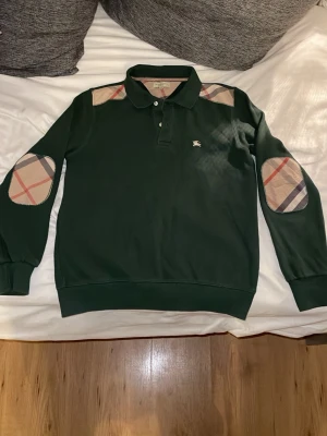 Grön stickad tröja från Burberry - Mörkgrön stickad tröja från Burberry med klassiska rutiga detaljer på armbågar och axlar. Tröjan har polokrage med knappar och en liten broderad logga på bröstet. Perfekt för dig som gillar stilrena och tidlösa plagg.