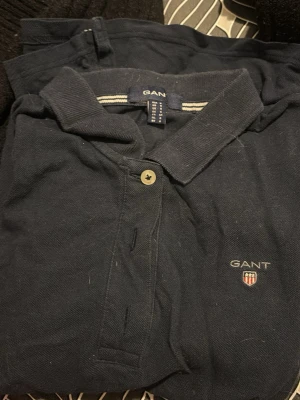 Marinblå pikétröja från GANT - Klassisk marinblå pikétröja från GANT med krage, knappslå och broderad logga på bröstet. Tillverkad i bomull och har korta ärmar. Perfekt för en stilren och avslappnad look.