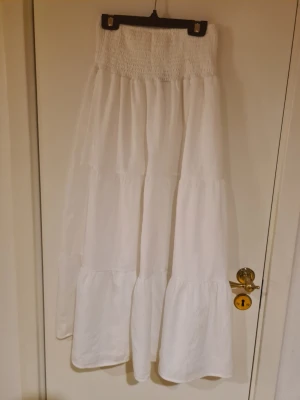 Vit långkjol med volanger - Säljer en vit långkjol med bred smock i midjan och flera volanglager. Kjolen har en luftig och flowy känsla, perfekt för varma dagar. Materialet känns mjukt och lätt, troligen bomull eller linne. Enkel och stilren design som är lätt att matcha. Fläckar förekommer 