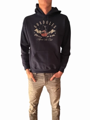 Svart hoodie Gondolen Alpine Ski Club - Svart hoodie från Gondolen med tryck framtill som visar en bergskabin och texten 'Alpine Ski Club'. Klassisk känguruficka och huva med dragsko. Mjuk bomullsblandning och avslappnad passform, perfekt för en chill look.