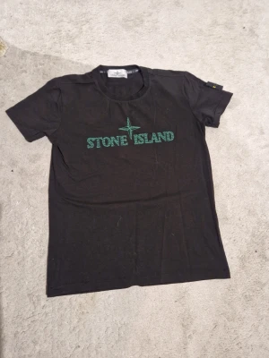 Svart Stone Island t-shirt - Svart t-shirt från Stone Island med grönt logotryck på bröstet och klassisk patch med kompass på ärmen. Rund halsringning och korta ärmar. Perfekt för dig som gillar streetwear och vill ha en stilren look.