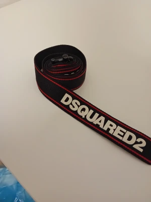 Svart DSQUARED2 textbälte - Snyggt svart bälte från DSQUARED2 med tydlig vit logotyp längs hela bandet och röda kanter. Bältet är gjort i slitstarkt textilmaterial och har ett metallspänne. Perfekt för att ge outfiten en sportig och trendig touch. Bältet har några år på nacken men är i bra skick. Passar på att köpa detta unika bälte från dsquared2 nu! 