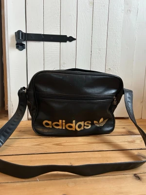 Svart axelväska från Adidas - Cool svart axelväska från Adidas med guldigt logotryck framtill. Väskan har justerbar axelrem, dragkedja och ett stort huvudfack samt ytterfack med dragkedja. Perfekt för att bära med sig det viktigaste i vardagen.