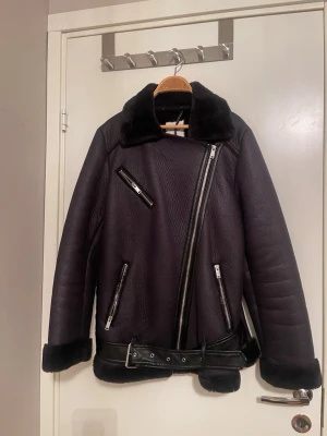 Svart pälsjacka med bälte och dragkedjor - ✨Snygg svart pälsjacka med markerad krage, bälte i midjan och flera silvriga dragkedjor. Jackan har långärmad design och mjuk päls på insidan samt vid ärmslut och krage. Perfekt för kalla dagar när du vill ha en cool look✨