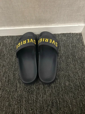 Svarta Sverige tofflor med gul text - Säljer ett par svarta tofflor med bred rem och gul text där det står 'SVERIGE' över foten. Tofflorna har platt sula och är tillverkade i syntetmaterial, perfekta för chill dagar hemma eller på stranden.