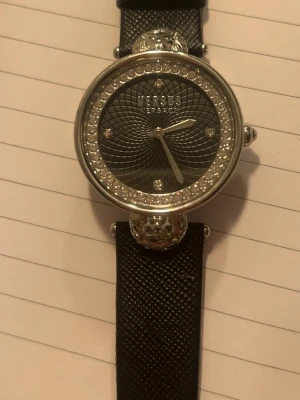 Versus Versace svart klocka med stenar - JÄTTA snygg klocka från Versus Versace med svart läderband och rund silverfärgad boett. Urtavlan är dekorerad med glittrande stenar runt kanten och har ett fint mönster priser kan diskuteras 💕 