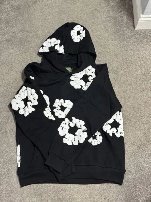 Svart hoodie med vita blommor - Säljer en svart hoodie med stora vita blommönster över hela tröjan. Hoodien har en klassisk huva och ribbade muddar vid ärmar och nederkant. Perfekt för dig som vill sticka ut med en cool och unik stil. Materialet känns mjukt och bekvämt.