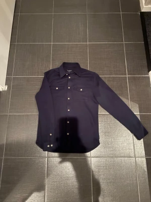 Mörkblå skjorta från Bondelid - Snygg mörkblå skjorta från Bondelid med klassisk krage och vita knappar. Skjortan har två bröstfickor med lock och är tillverkad i ett mjukt bomullstyg. Perfekt för en stilren och avslappnad look.