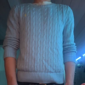 Ljusblå kabelstickad tröja Polo Ralph Lauren - Storlek L i barn, passar 155-170 cmLjusblå kabelstickad tröja från Polo Ralph Lauren med rund halsringning och klassisk gul broderad logga på bröstet. Tröjan har långa ärmar och ribbade muddar vid ärmslut och nederkant. Perfekt för en stilren och avslappnad look. Dock är det några få tal små brun fläckar men dom syns knappt