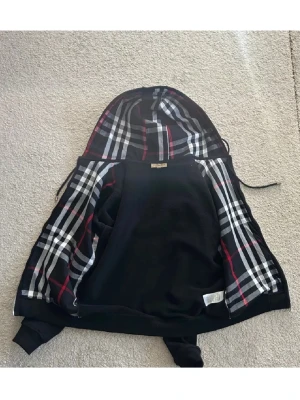 Burberry hoodie - Iprincip ny, använd 1-2 gånger!!                Svart hoodie från Burberry med dragkedja och klassiskt rutigt foder i huvan och insidan. Hoodien har ribbade muddar, fickor och justerbar dragsko i huvan. Materialet är mjukt och bekvämt, perfekt för en clean och stilren look. 
