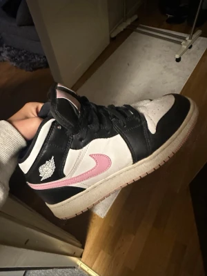 Nike Air Jordan 1 svart/vit/rosa - Snygga Nike Air Jordan 1 mid sneakers i svart, vitt och rosa. Skorna har klassisk svart snörning och rosa detaljer. Tillverkade i skinn (äkta). Perfekta för dig som vill sticka ut med stil.
