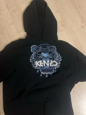 Svart Kenzo hoodie med tigertryck - Säljer en svart hoodie från Kenzo med det ikoniska tigerhuvudet och Kenzo Paris-loggan broderat i vitt och blått på bröstet. Hoodien har en stor huva och magficka framtill. Perfekt för dig som gillar streetwear och vill sticka ut.
