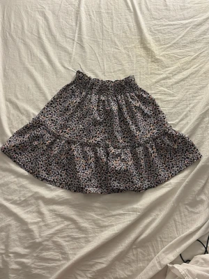 Blommig volangkjol från H&M - Supersöt blommig kjol från H&M i lila, svart och rosa toner. Kjolen har volang och resår i midjan för en bekväm passform. Perfekt för dig som gillar en lekfull och trendig stil.