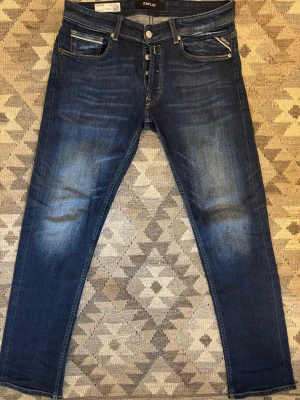 Blå Replay Grover jeans med knappar - Säljer ett par mörkblå Replay Grover jeans med snygg tvätt och knappar i gylfen. Klassisk femficksmodell med kontrastsömmar och läderpatch bak i midjan. Jeansen har en rak passform och är tillverkade i mjuk bomullsdenim.
