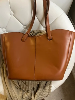 Brun shoppingväska i skinnimitation från ASOS  - Stilren shoppingväska i brun skinnimitation med två långa handtag. Väskan har en enkel och klassisk design med synlig söm i mitten och gott om plats för allt du behöver. Perfekt för dig som vill ha en snygg och praktisk väska till vardagen.