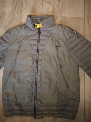 Grå dunjacka från Parajumpers - Snygg grå dunjacka från Parajumpers med hög krage och gul detalj vid halsen. Jackan har två stora bröstfickor med dragkedja och quiltad design. Perfekt för kalla dagar och har en modern, sportig look.