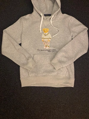Grå hoodie Polo Bear Ralph Lauren - Grå hoodie från Ralph Lauren med ikoniska Polo Bear-trycket framtill. Hoodien har snörning i huvan och en klassisk känguruficka. Mjuk och skön bomullsblandning, perfekt för en avslappnad och stilren look.