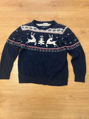 Stickad Jultröja - Mysig mörkblå stickad tröja från H&M med vitt och rött julmönster. Tröjan har motiv av renar, gran och snöflingor över bröstet och axlarna. Rund halsringning och ribbade muddar vid ärmslut och nederkant. Perfekt för vinterns alla tillfällen!
