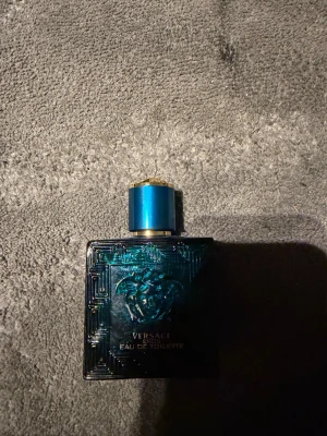 Versace Eros Eau de Toilette - Versace Eros Eau de Toilette i en stilren, fyrkantig flaska i metallic blått med Medusa-huvudet i relief på framsidan. Flaskan har ett elegant blått lock med gulddetaljer och är tillverkad i glas. En ikonisk doft som sticker ut i badrumshyllan.