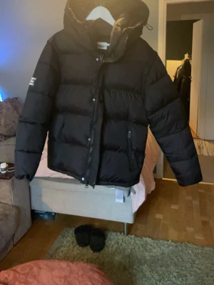 Svart pufferjacka från Burberry - Snygg svart pufferjacka från Burberry med huva och dragkedja framtill. Jackan har en quiltad design och praktiska fickor med dragkedja. Perfekt för kalla dagar när du vill hålla stilen och värmen. ( obs ja den kan bli till en väst med) storlek L men den passar M 