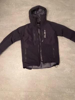 Sail racing jacka - Snygg svart dunjacka från Peak Performance med huva och flera dragkedjeförsedda fickor. Jackan har en modern passform och diskret logga på bröstet. Perfekt för kalla dagar och enkel att matcha med olika outfits.