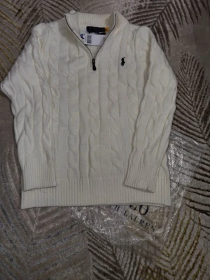 Vit kabelstickad tröja Polo Ralph Lauren - Kabelstickad vit tröja från Polo Ralph Lauren med halv dragkedja och broderad logga på bröstet. Tröjan har ribbade muddar och krage, perfekt för en clean och klassisk look. Materialet är mjukt och håller värmen.