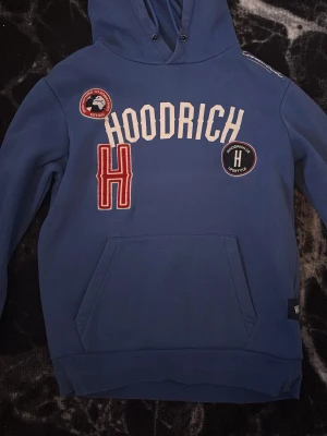 Blå hoodie från Hoodrich med patches - Blå hoodie från Hoodrich med stor vit logga på bröstet, röda och svarta patches samt en stor röd broderad H. Klassisk känguruficka framtill och justerbar huva med snören. Tillverkad i mjukt bomullsmaterial, perfekt för en avslappnad streetwear-look.
