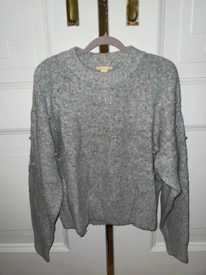 Grå stickad tröja med pärlor - Mysig grå stickad tröja med rund hals och dekorerad med pärlor och små glittriga stenar över hela framsidan och ärmarna. Tröjan har en avslappnad passform och ribbade muddar vid ärmslut och nederkant. Perfekt för dig som gillar detaljer och vill ha något extra.