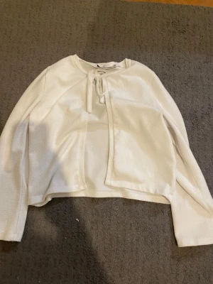 Vit fleecekofta med knytband - Mysig vit fleecekofta med långa ärmar och knytband framtill. Koftan har en rak passform och är tillverkad i mjukt fleecematerial som passar perfekt för kyliga dagar. Enkel och stilren design som är lätt att matcha med olika outfits.