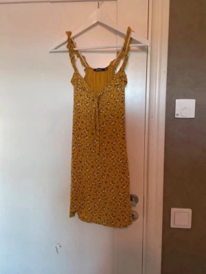 Gul blommig kortklänning Boohoo - Supersöt gul kortklänning från Boohoo med små vita blommor, volangdetaljer på axelbanden och knyt framtill. Perfekt för soliga dagar och har en lätt och luftig känsla. Axelbandsmodell och smickrande passform.