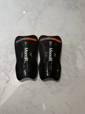 Monolit benskydd 14 cm kolfiber - Säljer ett par svarta benskydd från Monolit, 14 cm långa, tillverkade i kolfiber med snyggt mönstrad yta. De är lätta och smidiga, perfekta för dig som spelar fotboll och vill ha riktigt bra skydd utan att tumma på stilen.