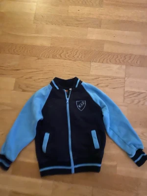 Blå tjocktröja från Djurgården DIF - Snygg tjocktröja från Djurgården i mörkblått med ljusblå ärmar och detaljer. Jackan har dragkedja framtill, ribbade muddar och klubbmärke på bröstet. Perfekt för dig som vill visa vilket lag du hejar på!