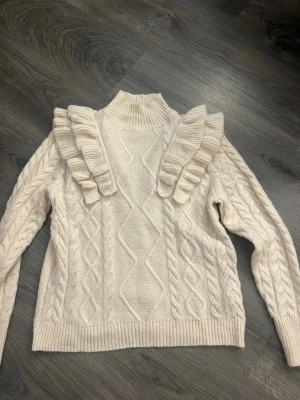Beige stickad tröja med volangdetaljer - Supersöt beige stickad tröja med höghals och unika volangdetaljer över axlarna. Tröjan har ett snyggt flätmönster framtill och ribbade muddar. Perfekt för dig som vill ha något extra till höst- och vinterlooken.
