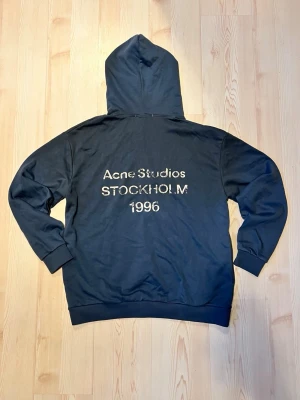 Mörkblå Acne Studios hoodie - Snygg mörkblå hoodie från Acne Studios med stor huva och tryck på ryggen där det står 'Acne Studios STOCKHOLM 1996' i vitt. Klassisk passform med ribbade muddar vid ärmslut och nederkant. Perfekt för dig som gillar Streetwear och vill ha något stilrent. 0bs lnte 0rglnal 