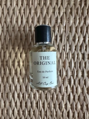 The Original Eau de Parfum 50 ml - En stilren parfymflaska från All Cap One med texten 'The Original'. Flaskan är genomskinlig med svart plastlock och vit etikett. Innehåller 50 ml Eau de Parfum. Enkel och modern design som passar perfekt på hyllan. Passar både tjejer och killar, doft av vanilj. Aldrig använd!