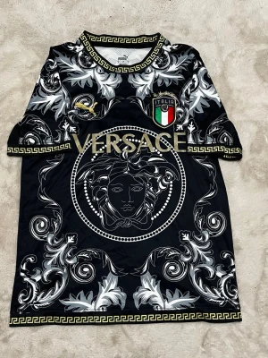 Svart och guldig fotbollströja Italien - Unik svart fotbollströja från VersaceXpuma med stora guldiga bokstäver, Medusa-motiv och detaljerade vita mönster. Italiens landslagsemblem på bröstet och klassiska grekiska bårder runt krage och ärmslut. Perfekt för dig som vill sticka ut på planen.