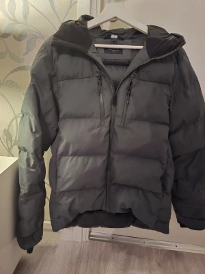 Grön dunjacka  - Säljer en svart dunjacka från Bik Bok med huva och två bröstfickor med dragkedja. Jackan har breda kanaler, hög krage och är perfekt för kalla vinterdagar. Enkel att matcha och riktigt varm.