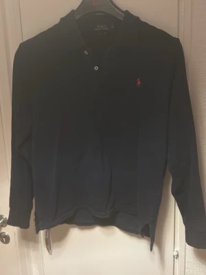 Mörkblå långärmad skjorta Polo Ralph Lauren - Mörkblå långärmad skjorta från Polo Ralph Lauren med klassisk krage, knappar framtill och den ikoniska röda logotypen broderad på bröstet. Skjortan har en normal passform och är tillverkad i mjuk bomull.
