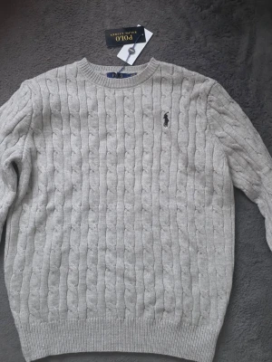 Ljusgrå kabelstickad tröja Polo Ralph Lauren - Säljer en ljusgrå kabelstickad tröja från Polo Ralph Lauren. Tröjan har rund halsringning, ribbade muddar och den klassiska broderade loggan på bröstet. Perfekt för dig som vill ha en clean och tidlös look.