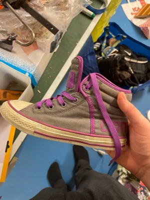 Converse All Star Sneaker - Säljer ett par höga Converse All Star sneakers i grått tyg med lila detaljer och snörning. Skorna har klassisk vit gummisula med lila rand och svart mönstrad undersida. Perfekta för dig som vill sticka ut med färgade detaljer. Pris går att diskutera