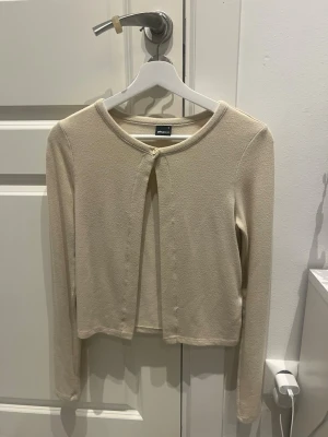 Beige  kofta  - Snygg beige c kofta  med rund halsringning och en knapp framtill. Koftan har långa ärmar och är gjord i ett mjukt, tunt material som passar perfekt till lager på lager. Enkel och stilren design som är lätt att matcha.