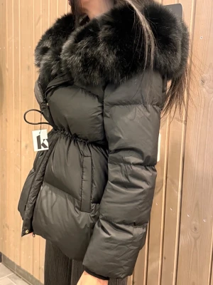 Svart dunjacka med stor pälskrage - Snygg svart dunjacka med stor fluffig pälskrage och markerad midja med snörning. Jackan har dragkedja framtill och quiltad design som ger en riktigt trendig look. Perfekt för kalla dagar när du vill vara både varm och stylish. 