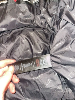 Svart dunjacka från Zavetti Canada - Snygg svart dunjacka från Zavetti Canada med diskret logotypmönster på fodret och klassisk röd lönnlövslapp på insidan. Jackan har en quiltad design och är perfekt för kalla dagar. Tillverkad i polyester för extra värme och komfort.