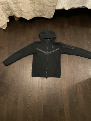 Svart Nike hoodie med dragkedja XS - Säljer en svart hoodie från Nike i storlek XS. Hoodien har hel dragkedja framtill, två fickor och en extra ficka på ärmen. Snygga grå detaljer över bröstet och klassisk Nike-logga. Tillverkad i mjukt material, perfekt för en sportig och avslappnad stil.