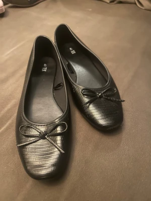 Svarta ballerinaskor med rosett H&M - Snygga svarta ballerinaskor från H&M med diskret strukturmönster och söt rosettdetalj framtill. Skorna har rund tå och platt sula, perfekta till många olika outfits. Klassisk och enkel design som passar till det mesta.