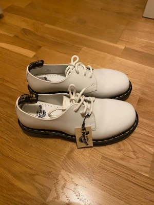 Vita Dr. Martens i skinn med snörning - Säljer ett par vita Dr. Martens med klassisk gul söm och snörning. Skorna är i skinn och har rund tå samt platt sula. Perfekta för dig som vill ha en clean och stilren look med edge.