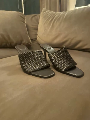 Svarta flätade pumps med klack - Stilrena svarta pumps med fyrkantig tå och hög klack. Skorna har en bred, flätad rem över foten som ger en trendig look. Perfekta för dig som gillar en modern och elegant stil. Materialet är skinnliknande och färgen är klassiskt svart.