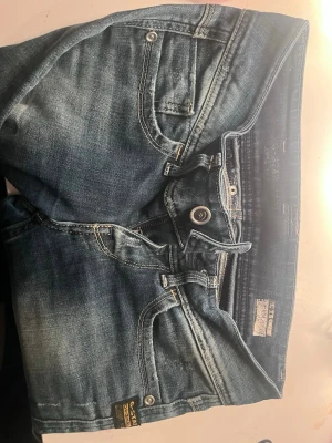 Blå G-Star RAW jeans med slitningar - Snygga blå jeans från G-Star RAW med klassisk femficksdesign och coola slitningar. Jeansen har en ljus tvätt och detaljer som kontrastsömmar och patch bak i midjan. Perfekta för en avslappnad och trendig look.